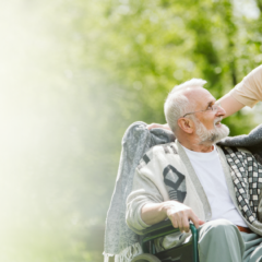 Caregiver smiling while placing a blanket over an elderly man in a wheelchair, enjoying a peaceful moment outdoors together.